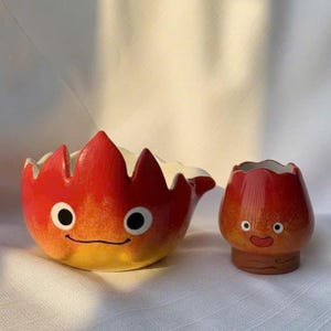May include: Two ceramic planters, one large and one small, in the shape of a fire demon. The large planter is bowl-shaped with a flame-like rim, and the small planter is a rounded cup. Both are red, orange, and yellow with black eyes and smiling faces.