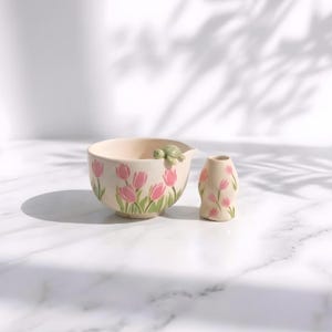 May include: A cream-colored ceramic bowl and a matching small vase, both decorated with pink tulip patterns and green stems. The bowl features a small, sculpted green turtle figurine perched on its rim, adding a unique detail to the set.