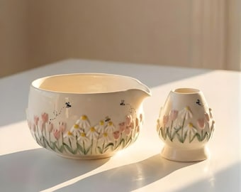 May include: Cream-colored ceramic bowl and small vase set. The bowl has a spout and is decorated with pink tulips, white daisies, green leaves, and black bees. The vase has a similar floral design.
