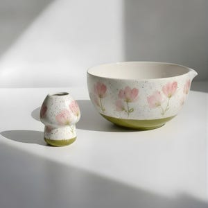 May include: A ceramic mushroom-shaped vase and a mixing bowl. The vase and bowl are white with pink floral designs and a speckled texture. The base of the vase and bowl are a light green color. The bowl has a spout.