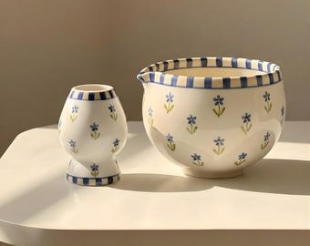 May include: Two ceramic pieces, a vase and a bowl, sit on a white surface. Both are white with blue floral designs and blue striped rims. The vase is egg-shaped, and the bowl has a spout.