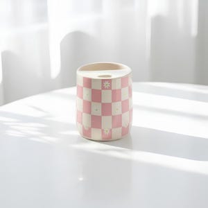 May include: A cylindrical ceramic toothbrush holder featuring a pink and white checkered pattern. Small white daisy flowers with yellow centers are printed within the squares of the pattern. The top of the holder has a single circular opening.