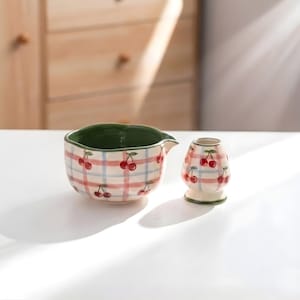 Puede incluir: Un cuenco de cerámica y un recipiente pequeño. Ambos son blancos con un patrón de cuadros rojos y azules e ilustraciones de cerezas rojas. El cuenco tiene un interior verde y un pico. El recipiente tiene forma redondeada.