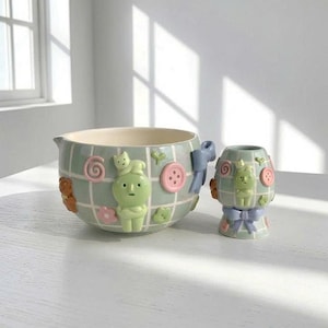 Puede incluir: Un cuenco de cerámica para mezclar y una taza más pequeña a juego, ambos con un patrón de cuadrícula verde claro. Los artículos están decorados con figuras en relieve de tonos pastel, incluyendo un personaje verde, un gato, botones rosas y lazos azules.