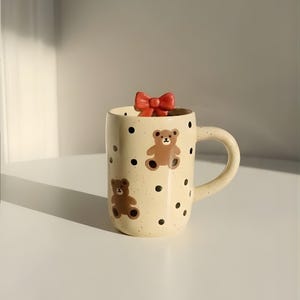 May include: A beige ceramic mug with a handle, decorated with brown teddy bears and black polka dots. A red bow sits atop the mug. The mug is on a white surface.