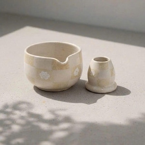 Könnte beinhalten: Eine cremefarbene Keramikschale mit Ausguss und eine kleine, passende Vase. Beide Teile weisen ein kariertes Muster mit kleinen weißen Blumenakzenten auf. Die Schale hat einen Durchmesser von etwa 10 cm, die Vase ist etwa 5 cm hoch.
