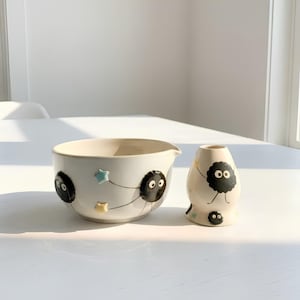 May include: A white ceramic mixing bowl and a matching small vase, both decorated with black fuzzy soot sprite characters with large white eyes. The bowl features small pastel-colored stars, while the vase shows a soot sprite with thin arms.
