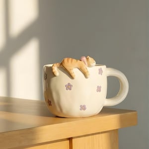 Op de afbeelding: Crèmekleurige keramische mok met een slapende oranje tabby kat figuur op de rand. De mok is versierd met paarse bloempatronen. De mok heeft een grote handgreep en staat op een houten oppervlak.