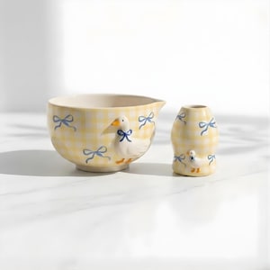Puede incluir: Un juego de cuenco y jarrón de cerámica amarilla con un estampado de cuadros y detalles de lazo azul. Cada pieza presenta un pato blanco con un lazo azul. El cuenco tiene un pico. El juego es para cocina o decoración del hogar.