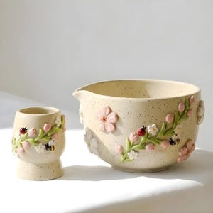 Puede incluir: Un cuenco de cerámica color crema con pico vertedor y una taza pequeña a juego, ambos con superficies moteadas y decoraciones en relieve 3D de flores de cerezo rosas, enredaderas verdes, pequeñas flores blancas, una mariquita y un abejorro.