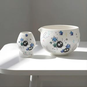 May include: White ceramic bowl and small vase set. Both pieces are decorated with a whimsical design featuring black, round characters with large eyes, blue flowers, and star accents. The bowl has a spout and the vase is small and rounded.