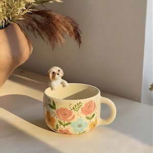 Puede incluir: Taza de cerámica color crema con un diseño floral en rosa, azul y amarillo. Una pequeña figura de perro blanco está posada en el borde. La taza tiene un asa redondeada y está llena de un líquido claro.