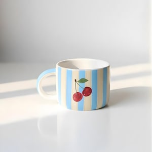 Puede incluir: Una taza de cerámica con interior blanco, asa azul claro y rayas verticales en azul claro y crema. La taza presenta un diseño pintado de dos cerezas rojas con una hoja verde.