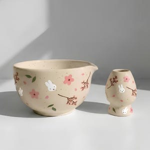 May include: A cream-colored ceramic mixing bowl with a pouring spout and a matching whisk holder. Both pieces feature a speckled finish decorated with small white rabbit faces, pink cherry blossom flowers, green leaves, and brown branches.