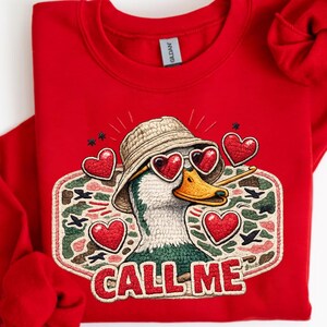May include: A red sweatshirt featuring a duck wearing heart-shaped sunglasses and a hat. The duck is inside a camouflage-style patch with the words "CALL ME". The design is a printed illustration, not embroidery.