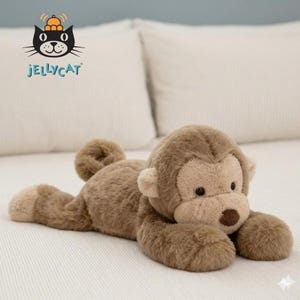May include: A plush, brown monkey toy with a light brown face, ears, and paws. The monkey is lying down on a white surface. The Jellycat logo is in the upper left corner.