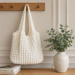 May include: A cream-colored, textured tote bag with long handles hangs from a wooden rack. The bag has a woven, puffy design. A white vase with green foliage sits on a wooden surface.