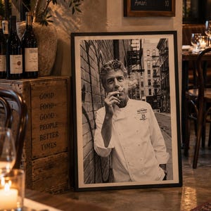 Peut inclure: Une photographie encadrée en noir et blanc d'Anthony Bourdain portant une veste de cuisine blanche, appuyé contre un mur de briques en fumant. L'impression présente le logo Les Halles sur la poitrine et est dans un cadre noir simple au sol.
