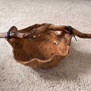 Pode incluir: Uma tigela rústica artesanal com uma forma orgânica. A tigela é marrom quente e terrosa com uma superfície texturizada. Um galho de madeira natural e curvo é preso no topo com barbante preto, adicionando um elemento artístico natural.