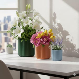 May include: Three decorative vases with flowers on a white table. The vases are green, orange, and blue, with vertical ribbed detailing. The flowers include white lilies, pink peonies, yellow tulips, and lavender. A small succulent is in a white pot.