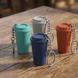 May include: Four miniature coffee cup keyrings in teal, grey, navy blue, and orange. Each cup has a matching lid and a silver chain with a keyring. The keyrings are arranged on a wooden surface.