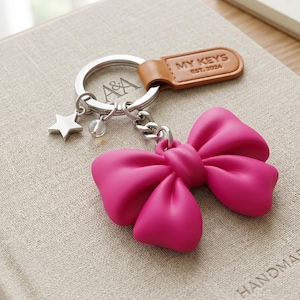 Puede incluir: Un llavero rosa en forma de lazo, con un llavero y una cadena plateados. El llavero tiene una etiqueta de cuero marrón que dice "MY KEYS EST. 2024". Una estrella plateada y una cuenta transparente cuelgan de la cadena.