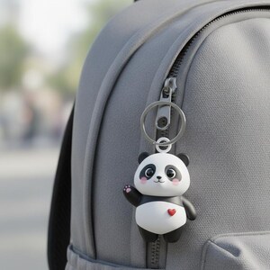 Op de afbeelding: Een panda sleutelhanger met een zilveren sleutelring bevestigd aan een grijze rugzak. De panda heeft zwart-witte vacht, een rood hart en zwaait. Een schattig accessoire.