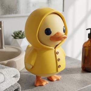 Könnte beinhalten: Gelbe Entenfigur mit Kapuzen-Regenmantel und drei Knöpfen. Die Ente hat schwarze Augen und einen orangefarbenen Schnabel und Füße. Die Figur steht auf einer grauen Oberfläche.