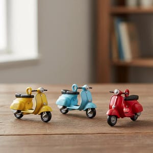 Puede incluir: Tres scooters en miniatura amarillos, celeste y rojos se exhiben sobre una superficie de madera. Cada scooter tiene un asiento y neumáticos negros, con detalles cromados. Los scooters están colocados en fila, mostrando su diseño clásico.