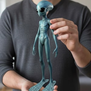 Puede incluir: Una figura de alienígena azul con grandes ojos negros y un físico esbelto. La figura tiene una cabeza grande, brazos y piernas largos y pies palmeados. El alienígena se sostiene frente a una persona que lleva una camiseta gris de manga larga.