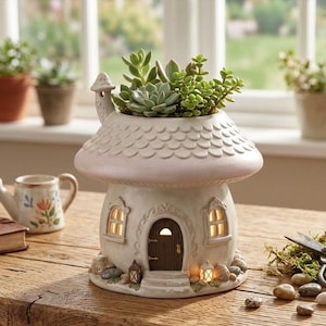 Op de afbeelding: Een grillige, paddenstoelvormige plantenbak met een lichte, getextureerde afwerking. De plantenbak is gevuld met groene vetplanten. Het huisvormige ontwerp heeft een deur, ramen en een schoorsteen. Andere items zijn een gieter, een boek en een schaar.