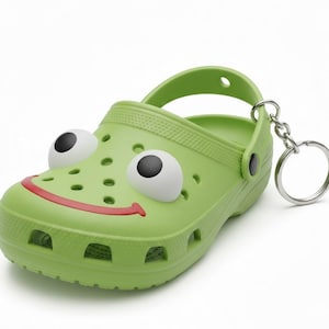 May include: A green frog-themed clog keychain with a smiling red mouth and large white and black eyes. The shoe has a strap and a silver keyring attached.