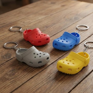 May include: Four miniature clogs keychains in red, blue, gray, and yellow are arranged on a wooden surface. Each clog has a silver keyring attached. The clogs are made of a solid material with multiple holes.