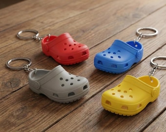 Op de afbeelding: Vier miniatuur klomp sleutelhangers in rood, blauw, grijs en geel liggen op een houten oppervlak. Elke klomp heeft een zilveren sleutelhanger. De klompen zijn gemaakt van een stevig materiaal met meerdere gaten.
