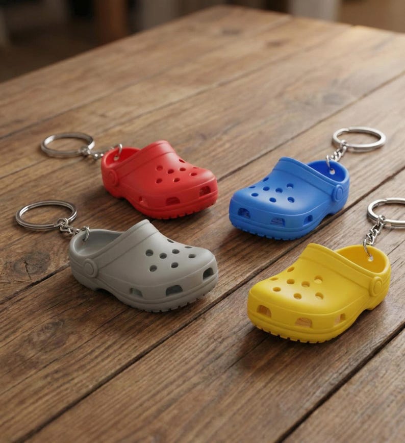 May include: Four miniature clogs keychains in red, blue, gray, and yellow are arranged on a wooden surface. Each clog has a silver keyring attached. The clogs are made of a solid material with multiple holes.