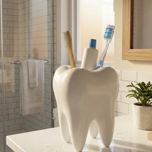 May include: A white, tooth-shaped toothbrush holder with three legs, containing a wooden toothbrush, a tube of toothpaste, and a blue and white toothbrush. The holder is on a white countertop in a bathroom.