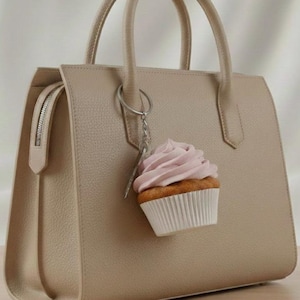 Puede incluir: Un bolso de mano beige con textura y un llavero de metal plateado. Un dije con forma de cupcake cuelga de la anilla, mostrando una base de color marrón dorado, un envoltorio de papel blanco y un remolino de glaseado rosa claro.