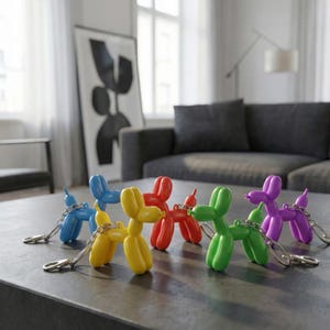 Puede incluir: Una colección de llaveros de perro globo de colores azul, amarillo, rojo, verde y morado. Cada perro tiene una cadena y un cierre de color plateado. Los llaveros se exhiben sobre una superficie gris oscuro.