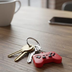 May include: A keyring with keys and a red video game controller charm. The charm has black buttons and a cross-shaped directional pad. The keys are gold and silver, on a wooden surface.