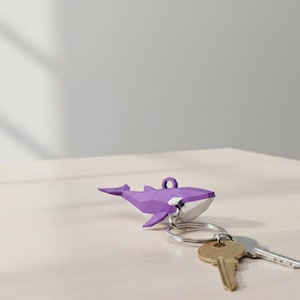 May include: A purple geometric whale keychain with a silver key ring and attached keys. The whale has a white belly and is resting on a light-colored surface. The keys are gold and silver.