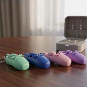 May include: Four colorful ballet slipper keychains in mint green, blue, pink, and purple are arranged on a wooden surface. A gray jewelry box is open in the background. Each slipper has a bow detail.