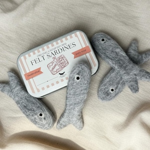 May include: A rectangular metal tin labeled Organic Catnip Infused Felt Sardines 100% Wool Handcrafted with a graphic of a sardine tin, accompanied by three grey felt fish toys, each featuring a single dark eye detail on a textured wool surface.