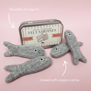 May include: Three grey felt fish toys infused with organic catnip, accompanied by a rectangular metal reusable storage tin. The tin label reads Organic Catnip Infused Felt Sardines, Handcrafted, 100% Wool, with a graphic of sardines.