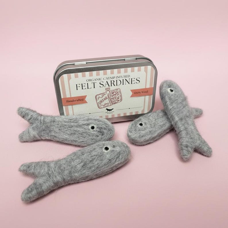 May include: A rectangular metal tin labeled Organic Catnip Infused Felt Sardines with three grey felt fish toys. The label features a graphic of a sardine tin and text including Handcrafted and 100% Wool. The fish have small black eyes.