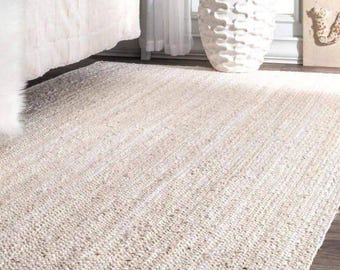May include: A rectangular, off-white rug with a woven texture. The rug is placed on a dark wood floor, with a bed and a decorative vase in the background. The rug's neutral color and simple design make it suitable for various interior styles.