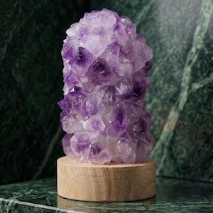 May include: A striking amethyst crystal cluster, displaying a rich purple colour, is set on a round, light-coloured wooden base. The crystal formation is dense and multifaceted, with visible crystalline structures. The background features a dark green marble surface.