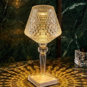 Puede incluir: Una lámpara de mesa transparente con un patrón de diamantes, base cuadrada y pantalla acampanada. La lámpara tiene apariencia de cristal y emite un brillo cálido. Es una lámpara decorativa.