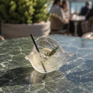 Könnte beinhalten: Eine klare, organisch geformte Glasschale mit gewellter Textur, die einen Cocktail mit Eis, einer Zitronenscheibe, einem Rosmarinzweig und einem schwarzen Strohhalm enthält, platziert auf einem grünen Marmortisch neben einem Weinglas und Oliven.
