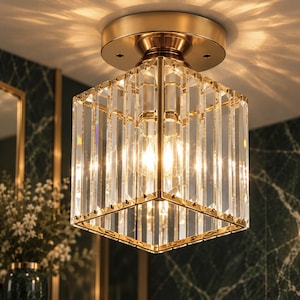 Könnte beinhalten: Eine quadratische Deckenleuchte mit einem goldfarbenen Metallrahmen und vertikalen, klaren Glasstäben. Die Leuchte strahlt ein warmes Licht aus und wirft durch die transparente Glasstruktur Lichtmuster an die Decke.