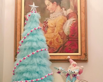 May include: A light blue tulle Christmas tree decorated with pink and white pom poms. The tree is topped with a silver star and is standing on a wooden table. A white sleigh with three reindeer figurines and a Santa Claus figurine is in the foreground.